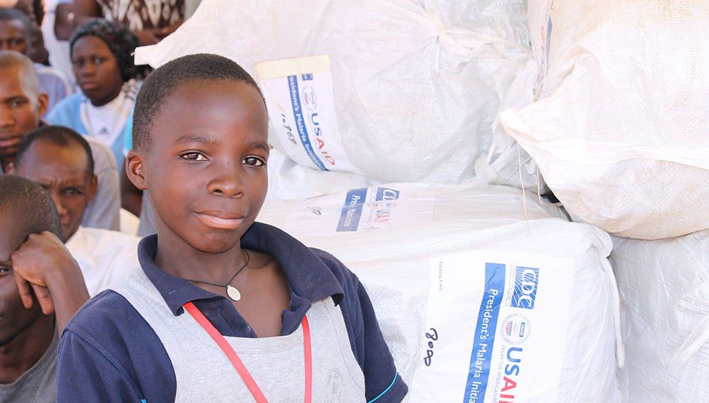 Boy stands next to stacks of nets at the end of project event for Uganda. Photo by, Stop Malaria Project in Uganda.